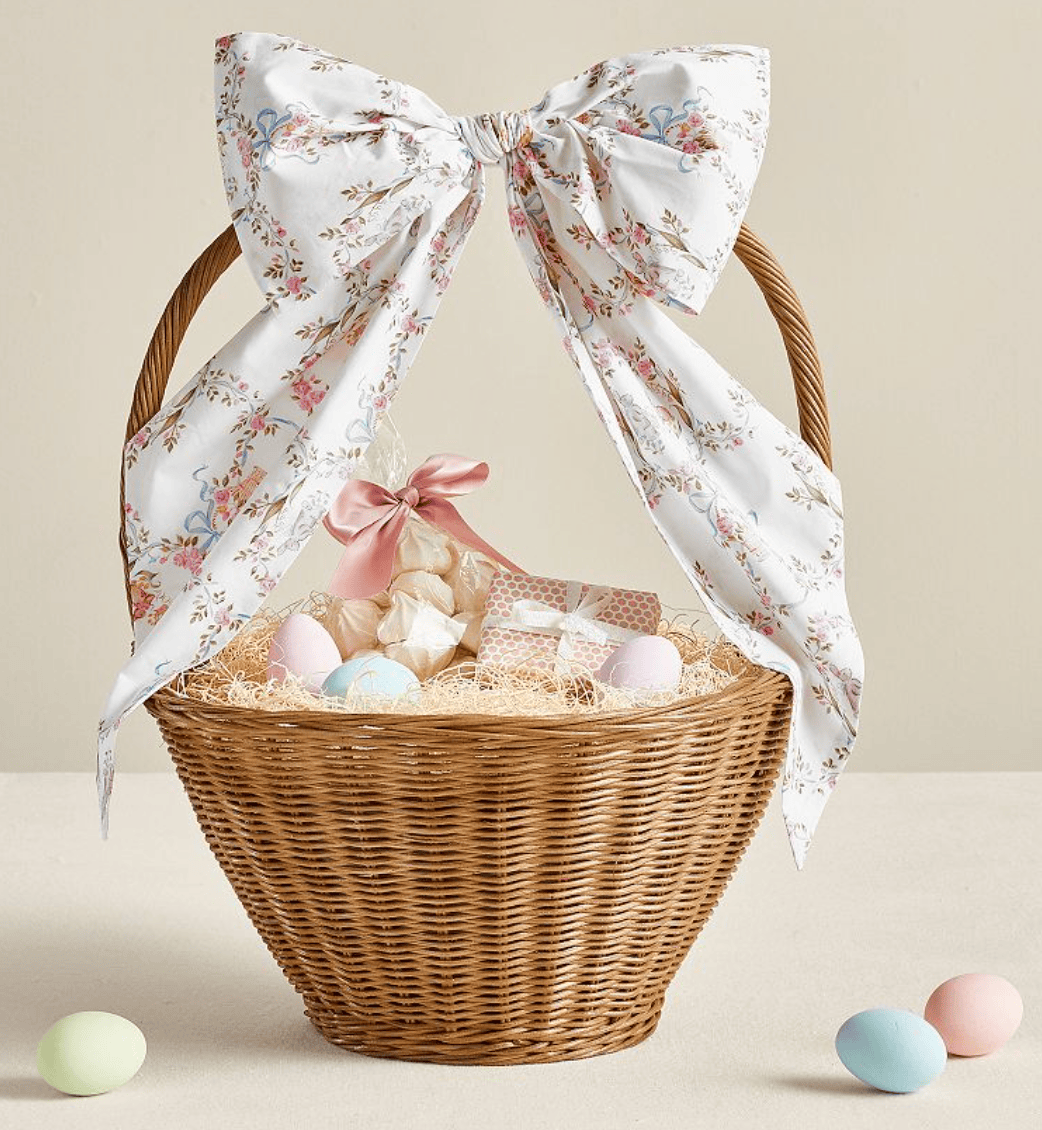 A wicker basket with a large floral bow on the handle, filled with pastel eggs, a fabric pouch tied with a pink ribbon, and small wrapped gifts—perfect for shopping with friends—sits on a light surface with three pastel eggs nearby.